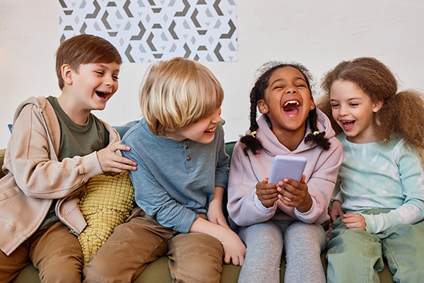 Group of cheerful little kids laughing emotionally and using smartphone indoors